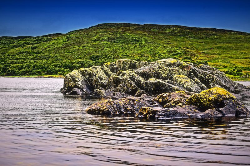Lichen-covered rocks on a shallow silver shore below a green hillside.