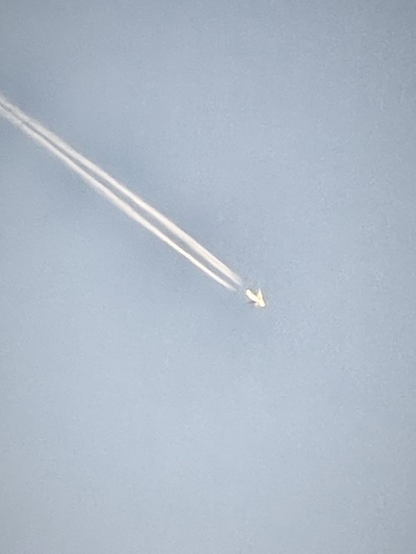 A high jet and its contrail cutting diagonally across a pale sky.