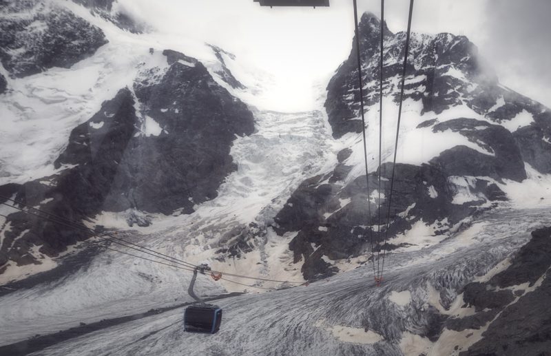 A cable car suspended above a snow-covered glacier and dark rock.
