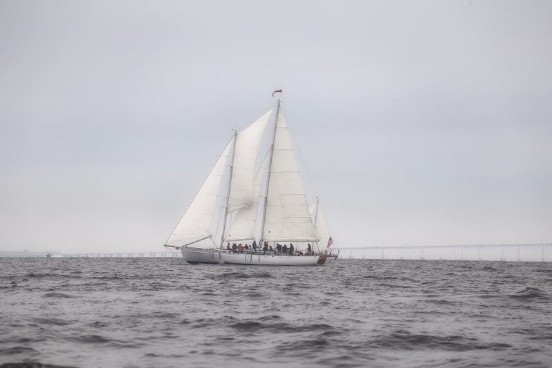 A two-masted schooner under full sail on a soft grey bay.