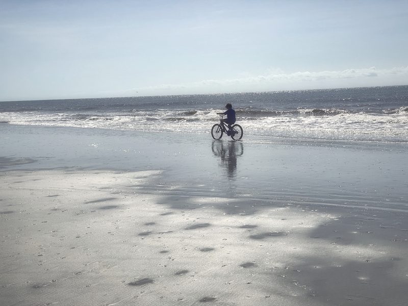 A lone cyclist on wet, reflective sand at the edge of the surf.