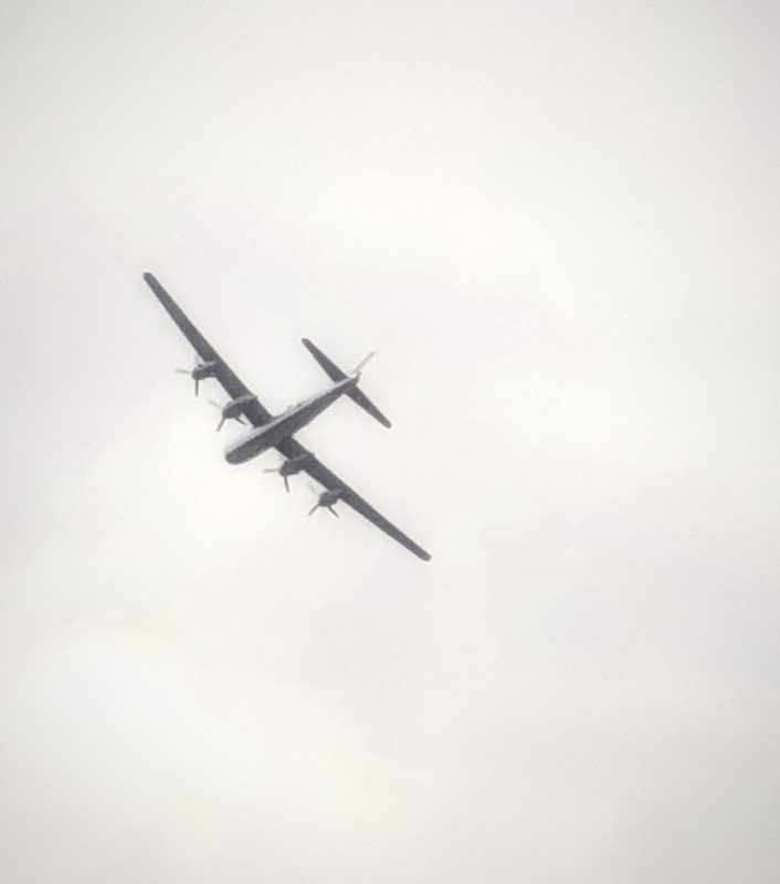 A four-engine warbird banking through a white, hazy sky.