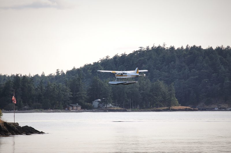 A floatplane lifting off a calm bay in front of a dark, forested shoreline.