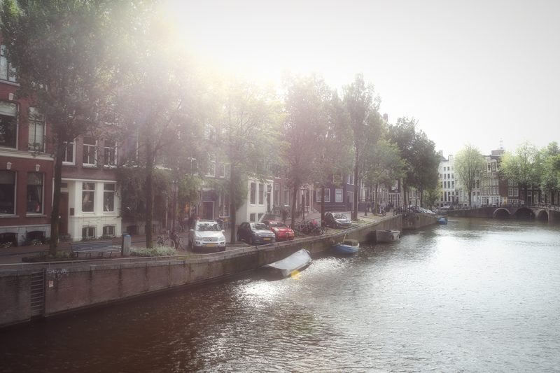 Hazy sun breaking through canal trees onto calm water and moored boats.