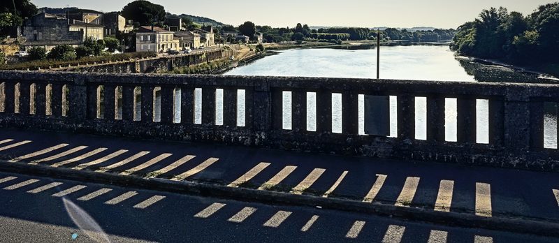 A stone bridge balustrade casting a row of striped shadows across the road above the Dordogne.