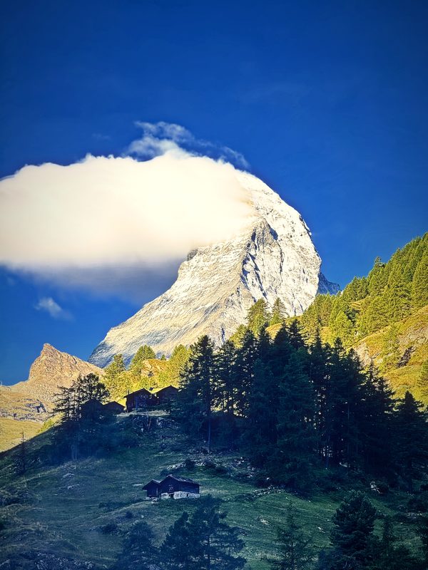 A dome of white cloud sitting on the Matterhorn above green larches and meadow.