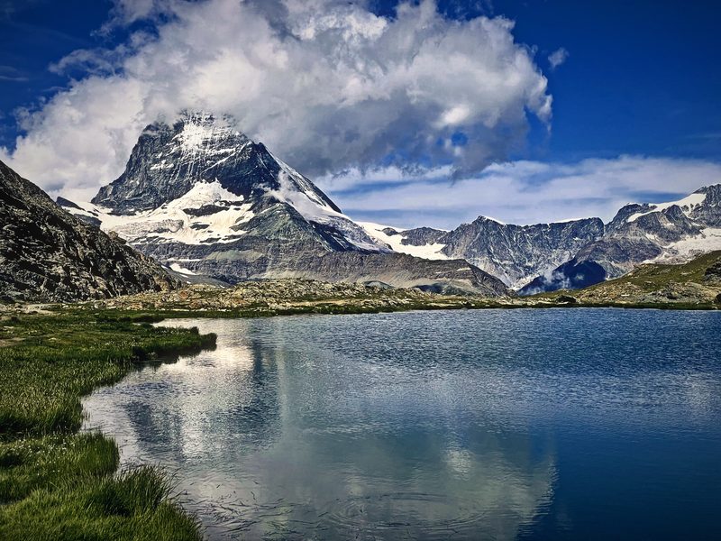 The Matterhorn and its clouds reflected in a still alpine lake edged with green meadow.