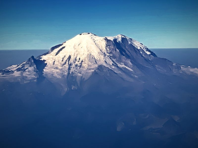 A broad snow-domed volcano rising above blue haze, seen from the air.