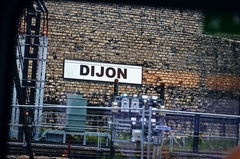 White station sign reading DIJON on a stone wall above tracks and equipment, seen from a train.