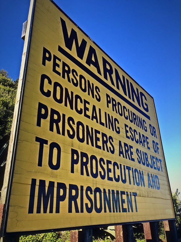 Yellow warning sign in heavy black capitals about concealing the escape of prisoners, against blue sky.