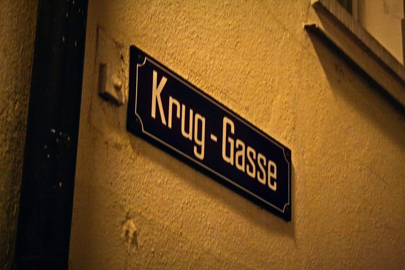 Dark blue Swiss street sign reading Krug-Gasse on a warm cream wall.