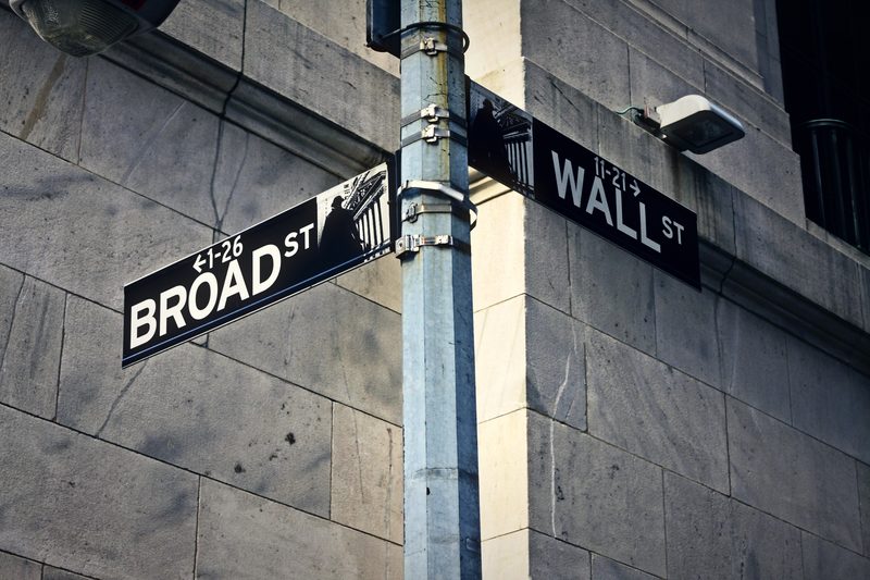 Two black street signs reading Broad St and Wall St on a lamppost against stone.