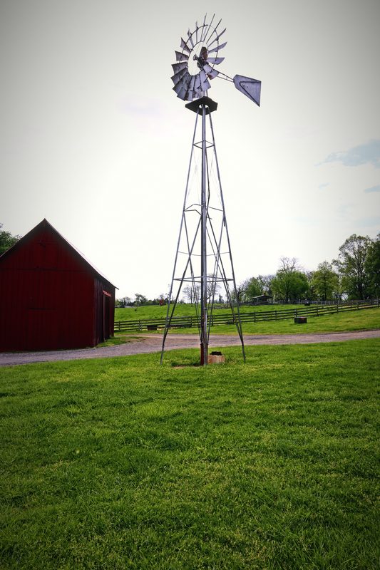 A metal windmill on a tall frame beside a small red barn on a green lawn.