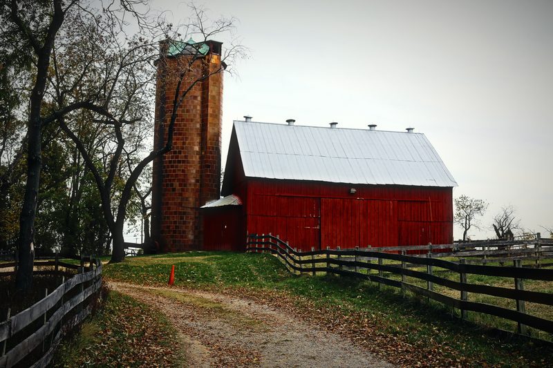 A red barn and brick silo at the end of a dirt lane with wooden fencing.