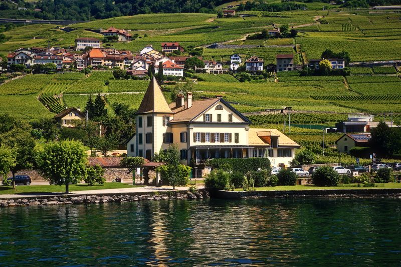 A house with a pointed yellow tower on the lakeshore below terraced vineyards.