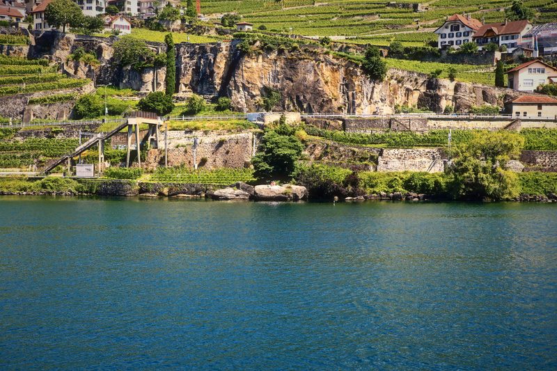 Rock cliffs and a village above vineyard terraces with a wide calm lake in front.
