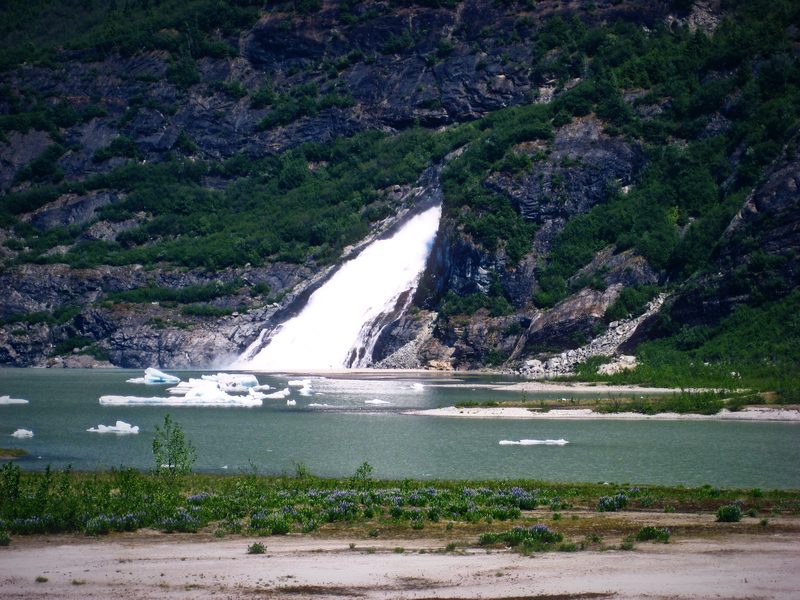 A white waterfall sliding down a dark mountainside into a pale glacial lake.
