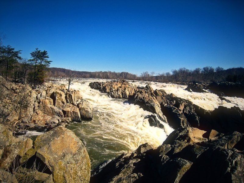 White rapids tearing through a wide field of broken rock under a deep blue sky.