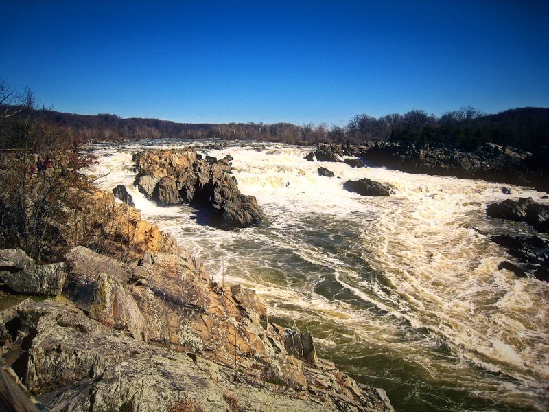 Churning white water spilling between jagged rocks under a clear blue sky.