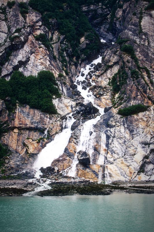 Waterfalls ribboning down a grey granite wall into green water.