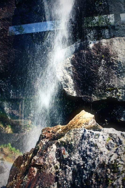 A tall waterfall falling behind a huge boulder in a rocky cove.