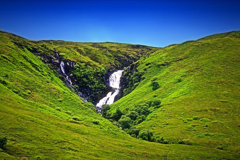 A waterfall dropping down a bright green glen under a deep blue sky.
