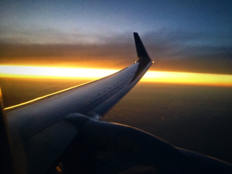 Silhouetted airplane wing pointing at a thin line of gold light between cloud and dusk.