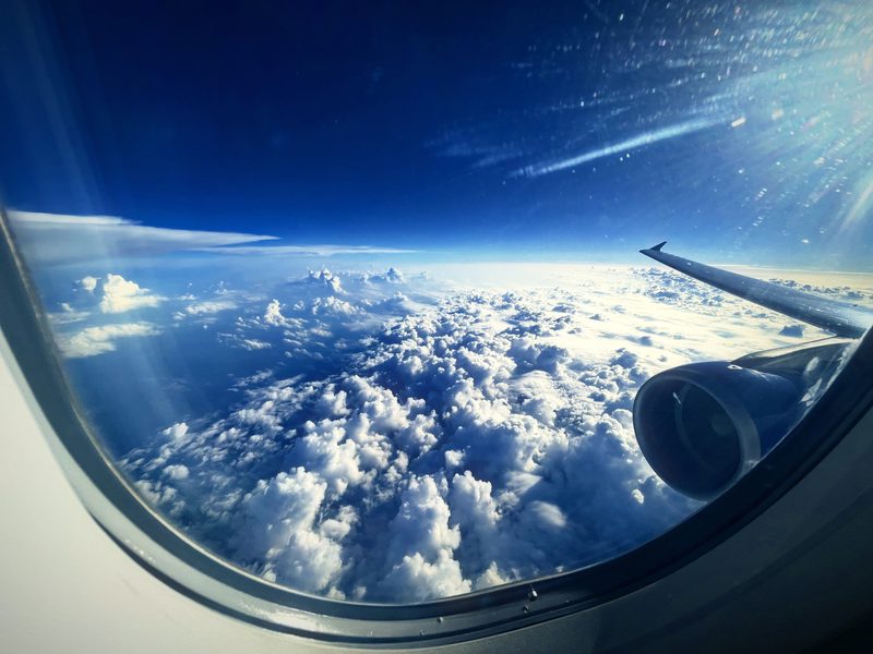 Curved airplane window framing towering cumulus clouds, an engine and deep blue sky.